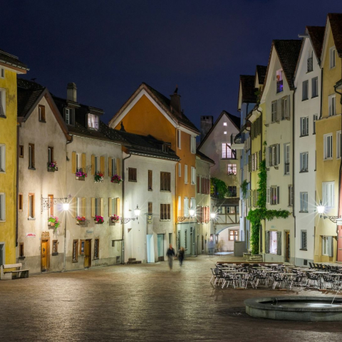Nächtlicher Blick über einen Platz in der Churer Altstadt mit beleuchteten, farbigen Häuserfassaden, Brunnen und leeren Restaurantterrassen.