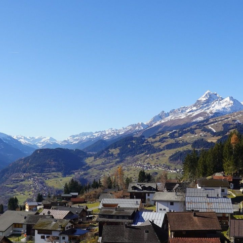 Weitblick über die Surselva mit einem Bergdorf und seiner Kirche im Vordergrund, bewaldeten Hängen sowie schneebedeckten Gipfeln am Horizont.