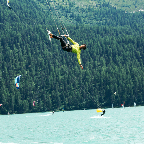 Wakesurfer in gelbem Outfit springt über türkisblauem Alpensee, dichte Fichtenwälder im Hintergrund, weitere Sportler im Wasser.