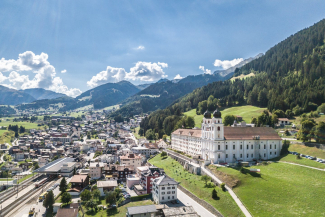 Luftaufnahme des barocken Klosters Disentis mit Blick über das sonnige Vorderrheintal in Richtung Sedrun und die bewaldeten Bündner Berge.