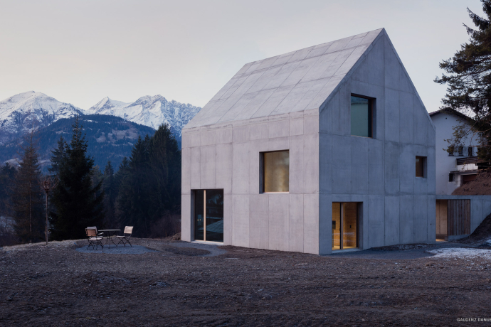 Modernes Betonhaus in Graubünden mit klaren Linien, grossen Fenstern und Terrasse vor einer verschneiten Berglandschaft und dunklen Tannen.