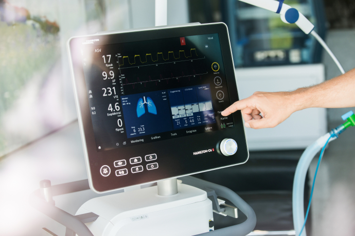 Eine medizinische Fachperson bedient den Touchscreen eines Beatmungsgeräts in einem Spitalzimmer und kontrolliert die angezeigten Atemwerte.