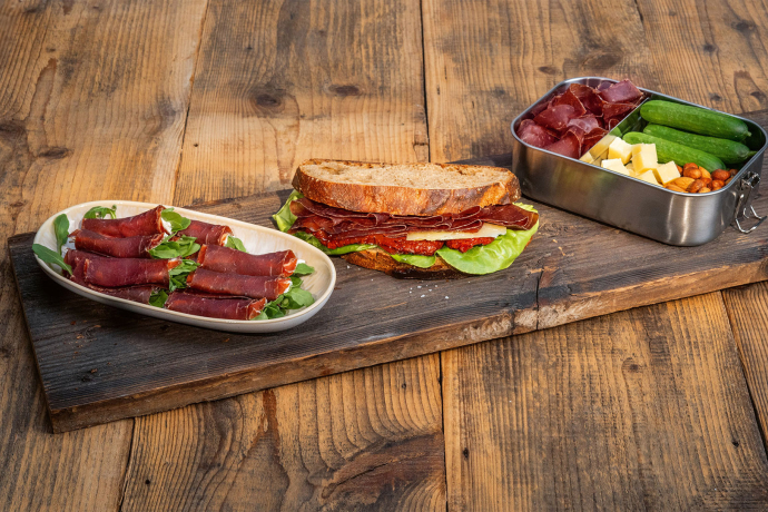 Bündnerfleisch-Plättli auf einem Holztisch mit Fleischröllchen, belegtem Brot sowie einer Box mit Käse, Nüssen und Gurken.