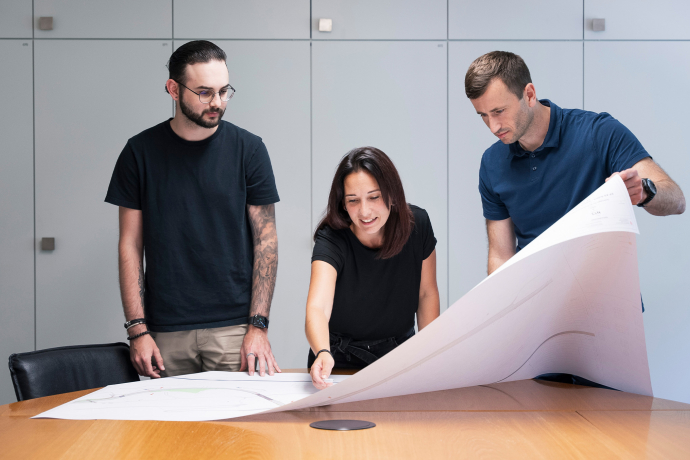 Drei Mitarbeitende besprechen gemeinsam eine grossformatige Planzeichnung an einem Besprechungstisch und stimmen Details für ein Projekt ab.