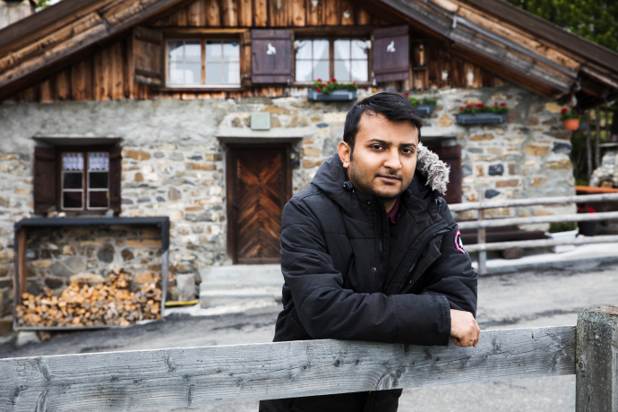 Person lehnt an einem Holzzaun vor einem traditionellen Berghaus mit Natursteinfassade und blickt direkt in die Kamera.