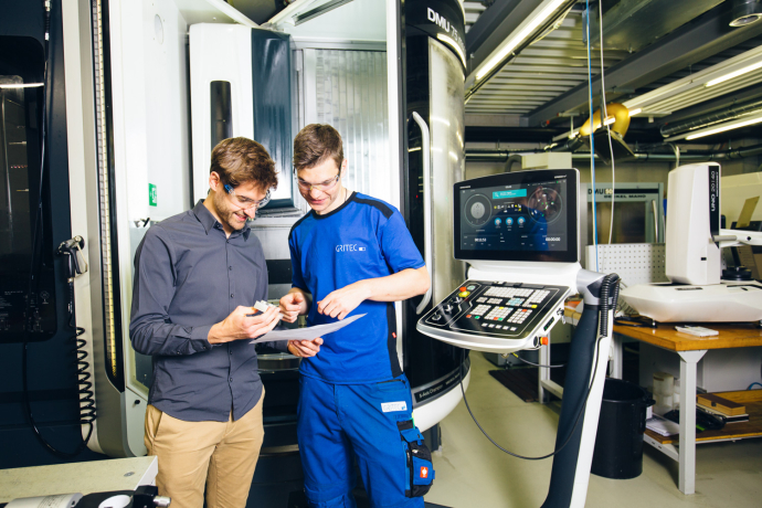 Zwei Mitarbeiter besprechen ein Werkstück und Unterlagen vor einer CNC-Maschine in einer modernen Produktionshalle.