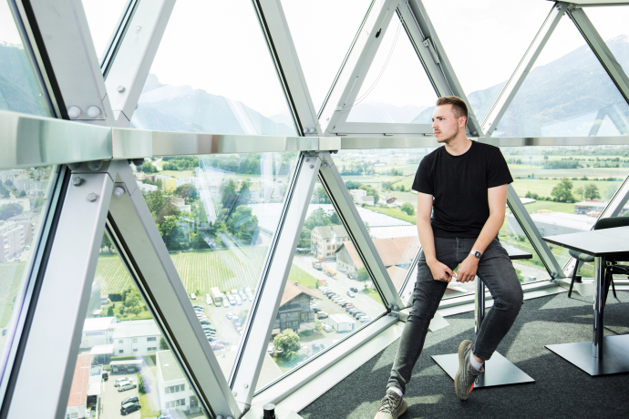 Mitarbeiter sitzt vor der grossen Glasfassade eines Aussichtsturms und blickt über das Tal mit Bergen, Feldern und Ortschaften im Hintergrund.