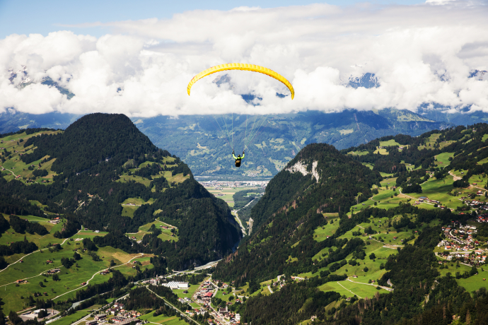 Gleitschirmflieger schwebt über der Berglandschaft Graubündens mit grünen Tälern, bewaldeten Hängen und Wolken im Hintergrund.