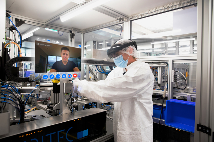 Mitarbeiter mit Schutzkleidung und Datenbrille arbeitet an einer Produktionsanlage, während ein Kollege per Videoanruf zugeschaltet ist.