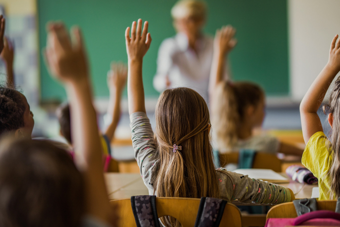 Schulkinder sitzen im Klassenzimmer und melden sich mit erhobenen Händen, während eine Lehrperson den Unterricht vor der Tafel gestaltet.