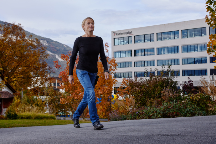 Eine Mitarbeitende des Kantonsspitals Graubünden geht durch eine herbstliche Gartenanlage vor dem modernen Spitalgebäude und der Berglandschaft.