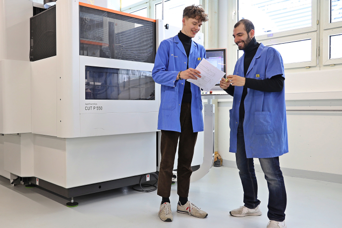 Zwei Mitarbeiter bei Polycontact besprechen technische Unterlagen in einer hellen Produktionshalle neben einer grossen industriellen Fertigungsmaschine.