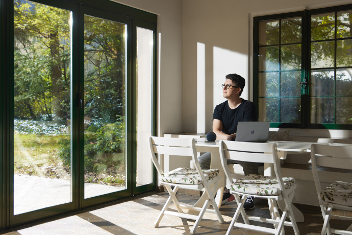 Mann mit Brille arbeitet am Laptop an hellem Schreibtisch in modernem Raum mit grossen Glasfenstern zur grünen Gartenlandschaft.