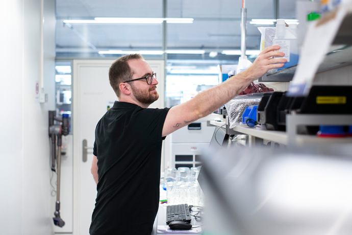 Ein Mitarbeiter bei Hamilton entnimmt in einem hellen Labor eine Probe aus einem Regal über einem Arbeitsplatz mit technischen Geräten.