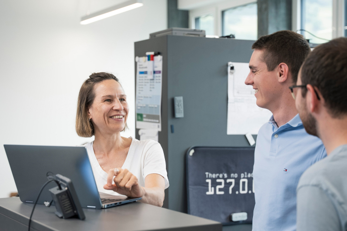 Drei Mitarbeitende bei INTEGRA unterhalten sich lächelnd an einem Stehpult mit Laptop in einem hellen, modernen Büro.