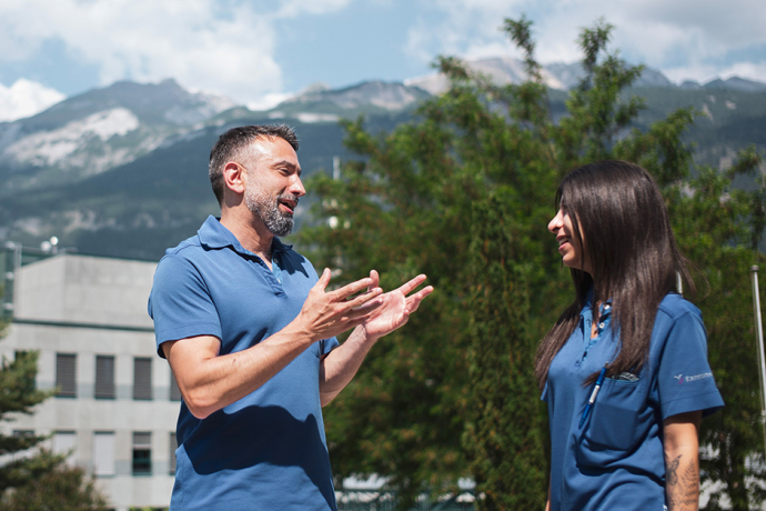 Zwei Mitarbeitende des Kantonsspitals Graubünden unterhalten sich im Freien vor dem Spitalgebäude und einer sommerlichen Berglandschaft.