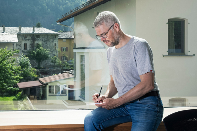 Mitarbeiter bei Repower sitzt am Fenster mit Blick auf alpines Dorf und schreibt Notizen, modernes Büro mit Berglandschaft im Hintergrund.