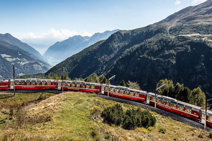 Der rote Bernina Express der Rhätischen Bahn fährt mit seinen Panoramawagen durch eine weitläufige alpine Berglandschaft.