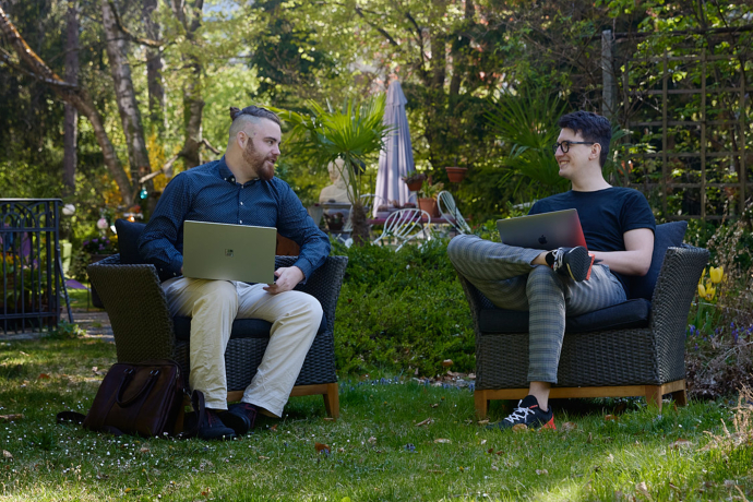 Zwei Männer sitzen mit Laptops in Korbsesseln im grünen Garten, umgeben von Bäumen und Pflanzen, diskutieren bei schönem Wetter.