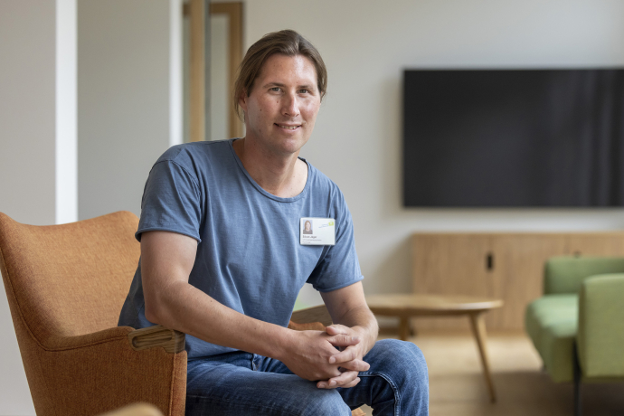 Mitarbeiter bei Psychiatrische Dienste Graubünden sitzt in modernem Wartezimmer mit braunem Sessel, grünem Sofa und Bildschirm im Hintergrund.