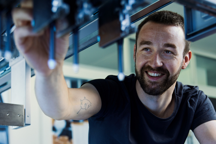 Ein lächelnder Mitarbeiter von HamiltonInfo montiert in einer modernen Werkhalle präzise Bauteile an einer technischen Anlage.
