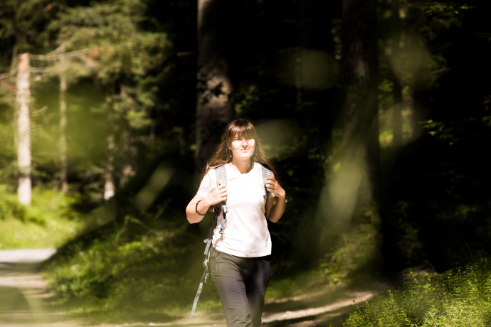 Eine Frau wandert mit Rucksack auf einem sonnendurchfluteten Waldweg durch einen dichten Wald mit hohen Bäumen.