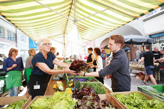 „Eine Markthändlerin übergibt einem Kunden frischen Salat an einem belebten Marktstand mit Gemüse unter einem gestreiften Sonnendach.