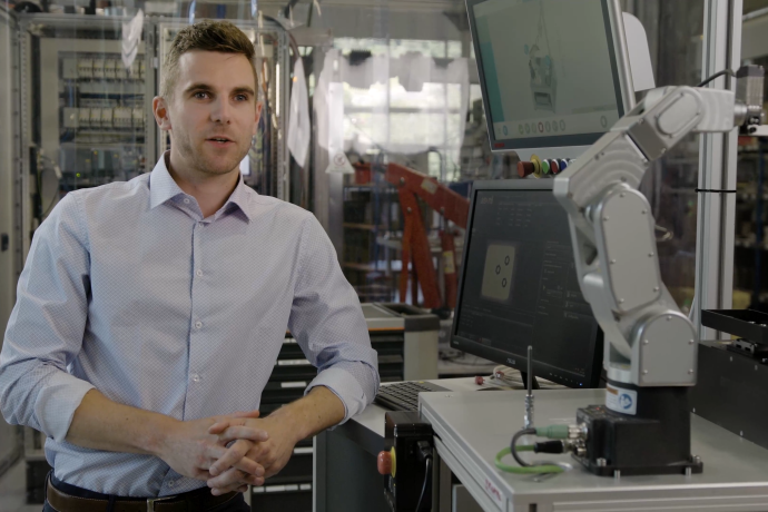 Mann an seinem Arbeitsplatz neben einem Roboterarm und Monitoren in einer modernen Produktions- und Entwicklungsumgebung.