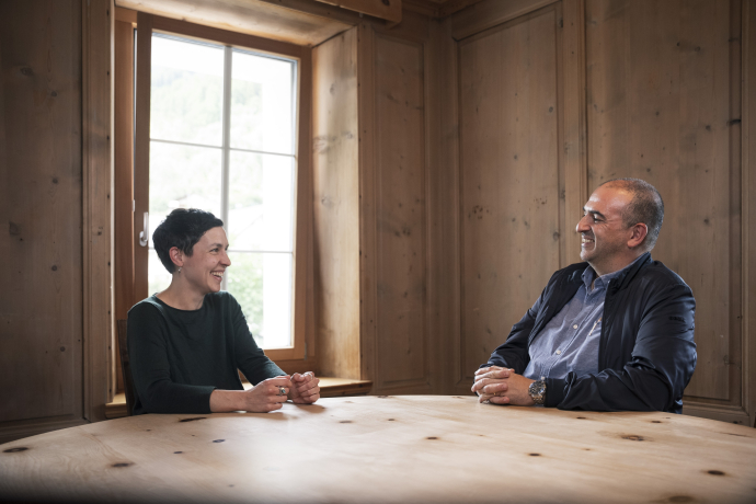 Two employees of Clinica Holistica Engiadina smile and chat at a wooden table in a cosy, wood-panelled room.