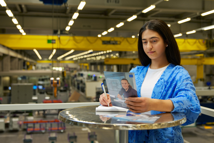 Eine Mitarbeiterin bei TRUMPF macht sich an einem Stehtisch Notizen zu einer Broschüre mit Blick auf die moderne Produktionshalle.