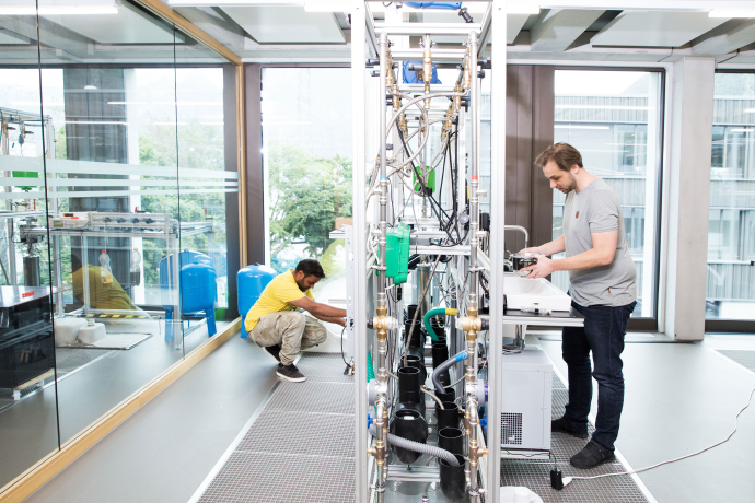 Modernes Laboratorium mit großen Fenstern: Zwei Techniker arbeiten an wissenschaftlichen Geräten und Versuchsaufbauten, helle Raumgestaltung mit Stadtblick.