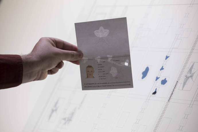 Eine Hand hält für ein Fenster in die Zukunft ein transparentes Ausweisdokument vor einen beleuchteten Bildschirm mit technischen Entwürfen.