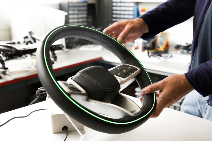 Modernes Lenkrad mit grünem LED-Ring für Fahren nach Farben wird mit beiden Händen gehalten, Touch-Steuerung und Werkstatt im Hintergrund.