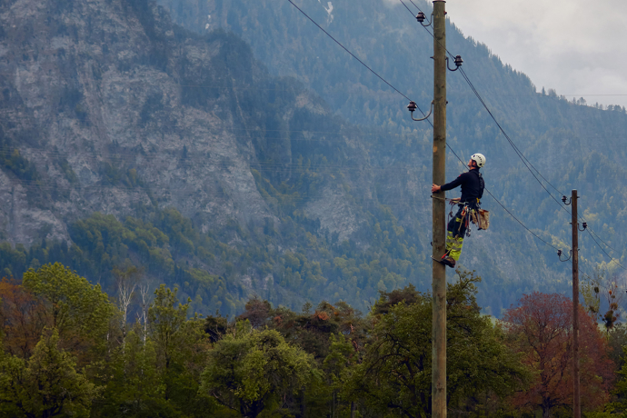 Ein Mitarbeiter von Repower klettert gesichert an einem Strommast und arbeitet an einer Leitung vor einer bewaldeten Berglandschaft.