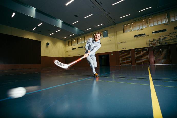 Ein Hockeyspieler schiesst mit einem Unihockeystock einen weissen Ball durch eine grosse, dunkel beleuchtete Sporthalle.