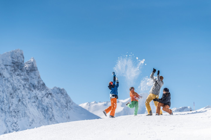 Eine Familie wirft auf einer verschneiten Piste jubelnd Schnee in die Luft, umgeben von eindrucksvollen Gipfeln unter blauem Winterhimmel.