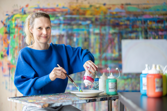 Frau bei Psychiatrische Dienste Graubünden malt mit Pinsel und Farben an Kunstwerk, bunte abstrakte Gemälde im Hintergrund, therapeutische Kunstaktivität.