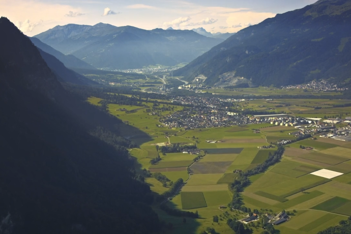 Panorama des Bündner Rheintals mit Feldern, einem Dorf und bewaldeten Berghängen vor der Kulisse der Alpen.