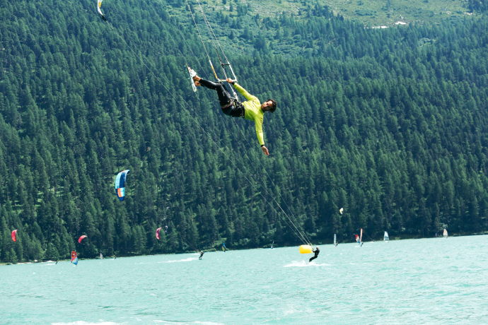Wakesurfer in gelbem Outfit springt über türkisblauem Alpensee, dichte Fichtenwälder im Hintergrund, weitere Sportler im Wasser.