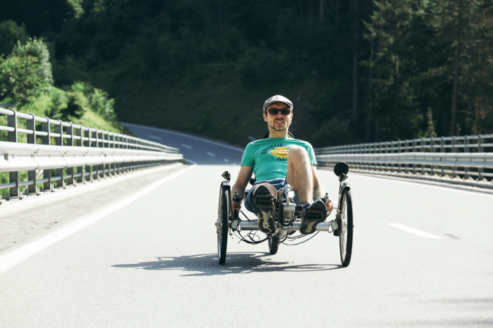 Ein Tüftler im Dienste der Natur: Mann in Liegerekumbent-Fahrrad auf breiter Brücke mit Metallgeländer, Wald im Hintergrund, sonniger Tag.