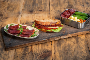 Bündnerfleisch-Plättli auf einem Holztisch mit Fleischröllchen, belegtem Brot sowie einer Box mit Käse, Nüssen und Gurken.