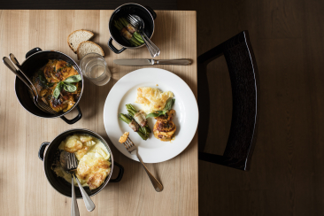 Alt-Text-Länge prüfen Stillleben mit einem angerichteten Teller, Kartoffelgratin, einem Gemüsegericht, Brot und Besteck auf einem gedeckten Holztisch.