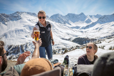 Eine Serviceangestellte reicht einem Gast auf einer sonnigen Bergterrasse ein Bier, vor der eindrucksvollen Kulisse verschneiter Alpengipfel.