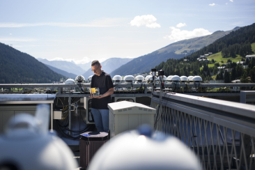 Ein Forscher misst auf einem Bergdach mit technischen Sensoren die Sonneneinstrahlung, während sich hinter ihm die Bündner Alpen erstrecken.