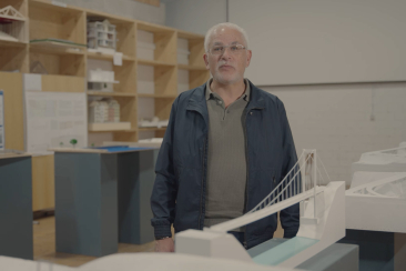 Ein Mann steht in einem Architekturatelier vor einem detailreichen Modell einer Hängebrücke, umgeben von weiteren Entwürfen und Modellen.