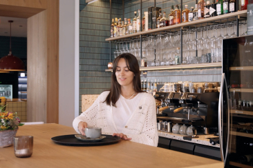 Eine Frau geniesst an der gemütlichen Bar eines modernen Cafés einen Kaffee, vor ihr eine Espressomaschine und ein Blumenstrauss.