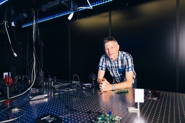 Ein Mann arbeitet in einer abgedunkelten Forschungsumgebung an einem optischen Messtisch mit Kabeln, Lasertechnik und elektronischen Geräten.