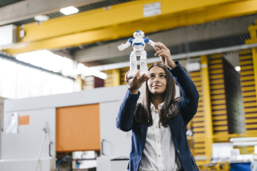 Frau hält einen kleinen humanoiden Roboter in den Händen und betrachtet ihn aufmerksam in einer modernen Produktionshalle.