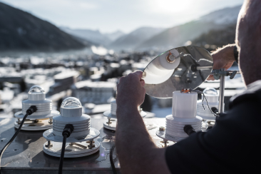 Ein Mitarbeiter justiert mehrere Messinstrumente auf einem Berg mit Blick über ein alpines Tal und die darunterliegende Ortschaft.