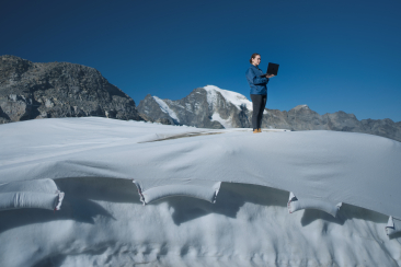 Eine Forscherin arbeitet mit einem Laptop auf einem Gletscher, umgeben von schneebedeckten Gipfeln und klarem, tiefblauem Himmel.
